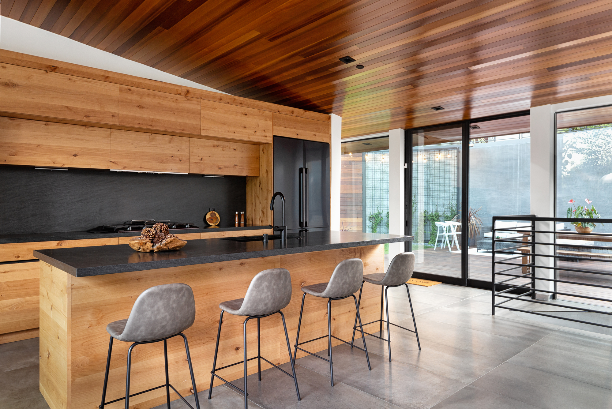 Modern kitchen with natural wood cabinetry, black countertops, a large island, and sliding glass doors opening to an outdoor living space. Modern kitchen with natural wood cabinetry, black countertops, a large island, and sliding glass doors opening to an outdoor living space.