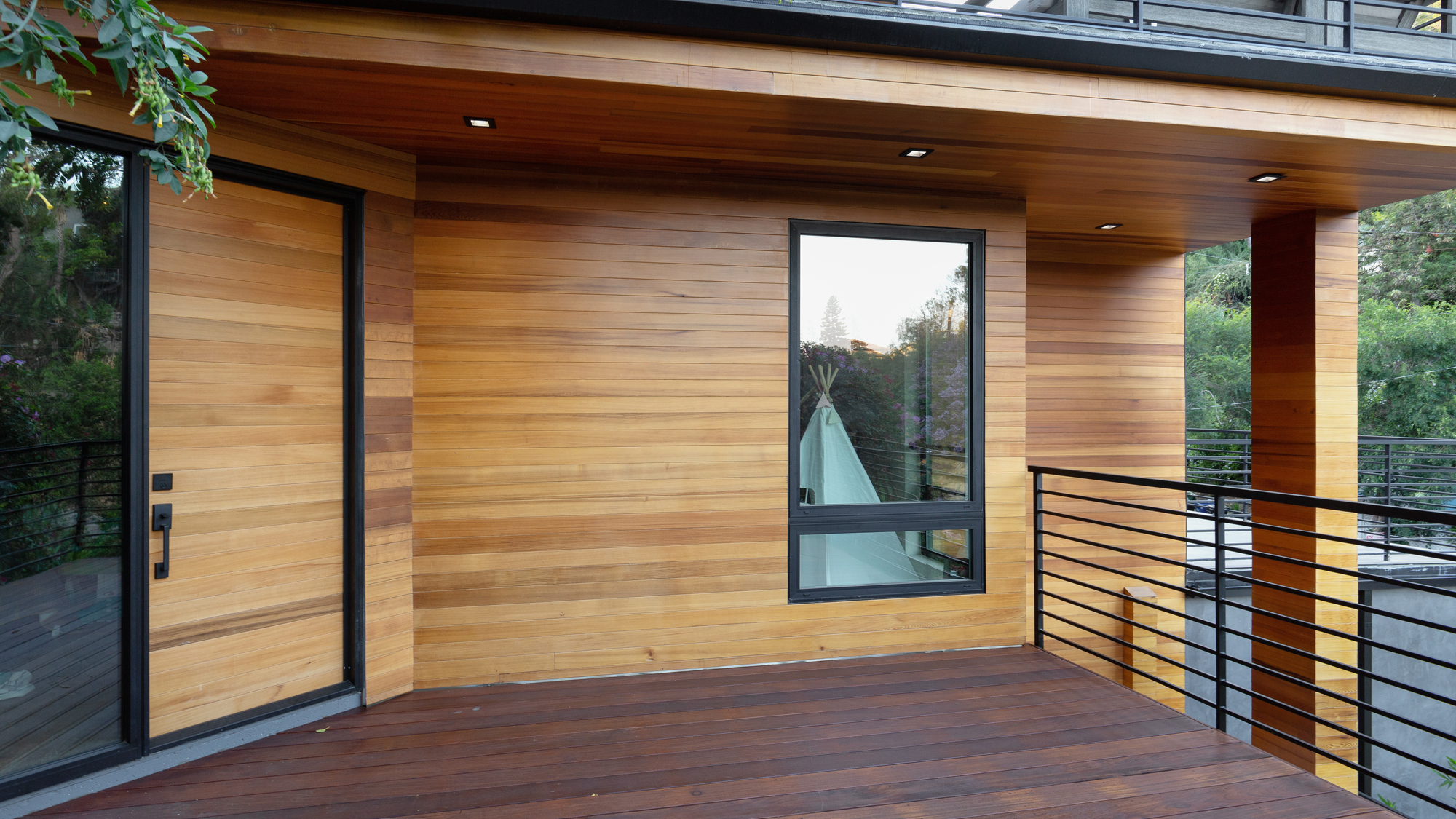 Covered outdoor living space with warm wood siding, wood decking, black-framed windows, and modern metal railings. Covered outdoor living space with warm wood siding, wood decking, black-framed windows, and modern metal railings.