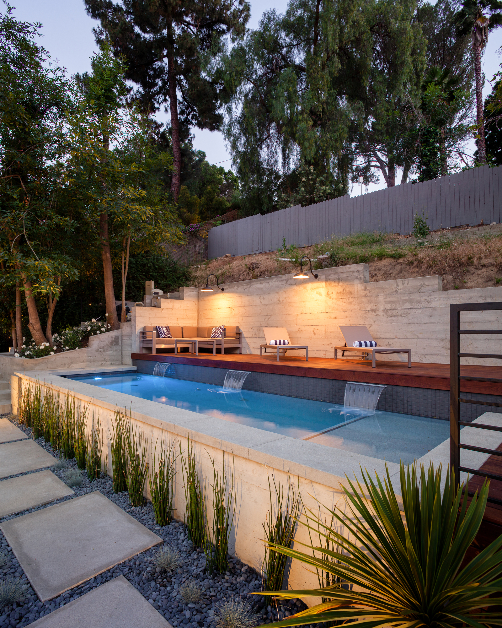 Modern hillside pool with waterfall features, wood decking, lounge seating, concrete retaining walls, and landscaped stepping-stone paths. Modern hillside pool with waterfall features, wood decking, lounge seating, concrete retaining walls, and landscaped stepping-stone paths.