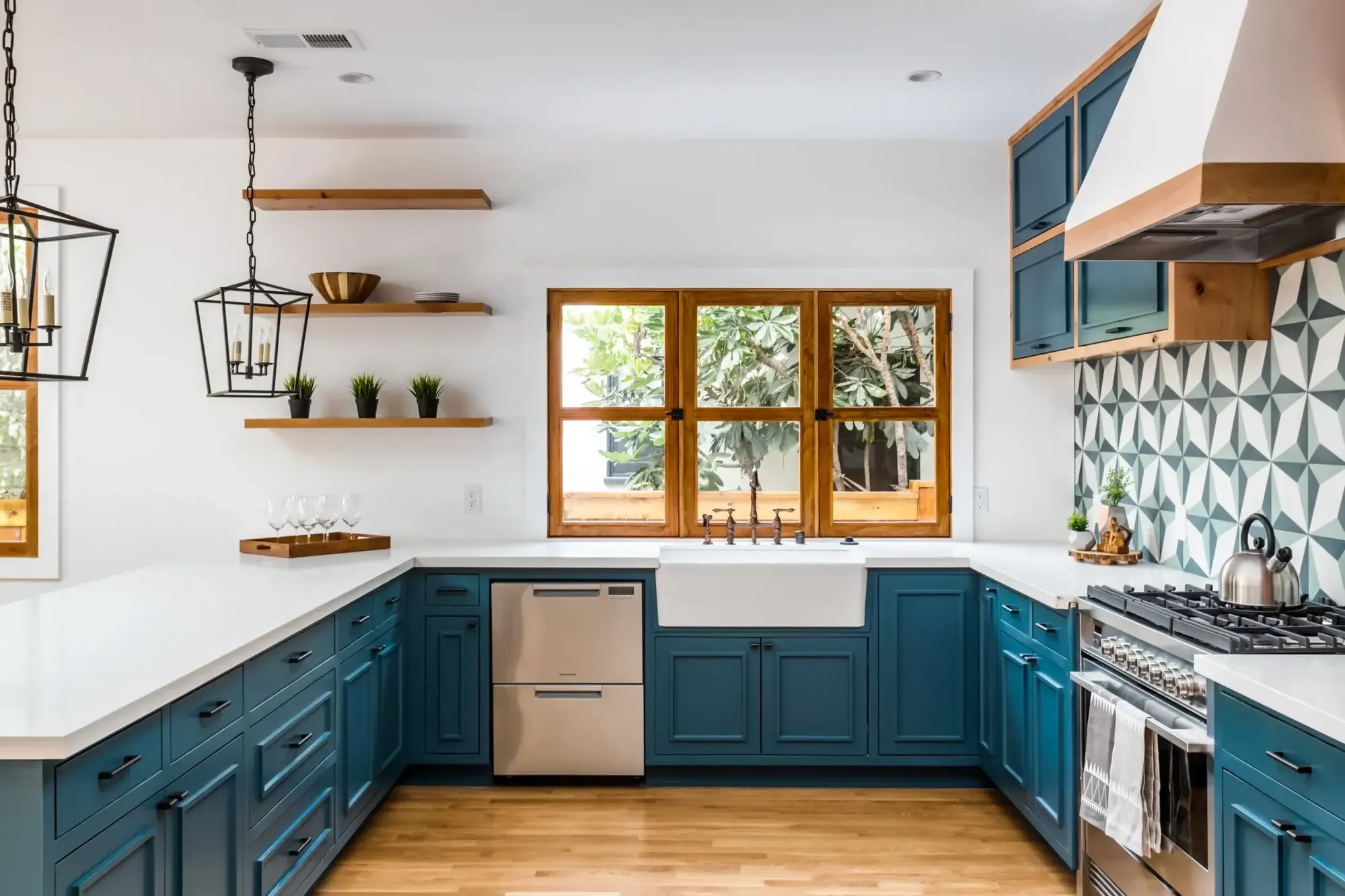 Teal kitchen cabinetry with white countertops, warm wood trim, geometric tile backsplash, farmhouse sink, and wood-framed windows.