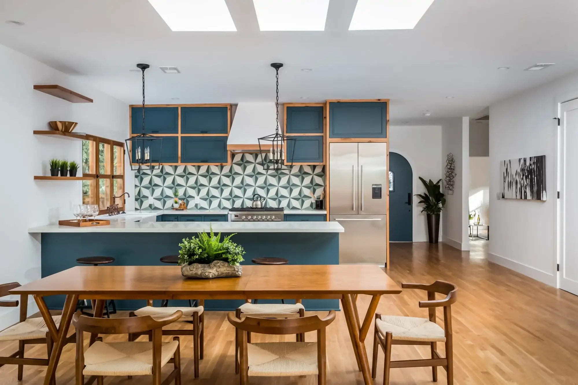 Open kitchen and dining area with teal cabinetry, geometric tile backsplash, wood accents, large peninsula, and skylights bringing in natural light.
