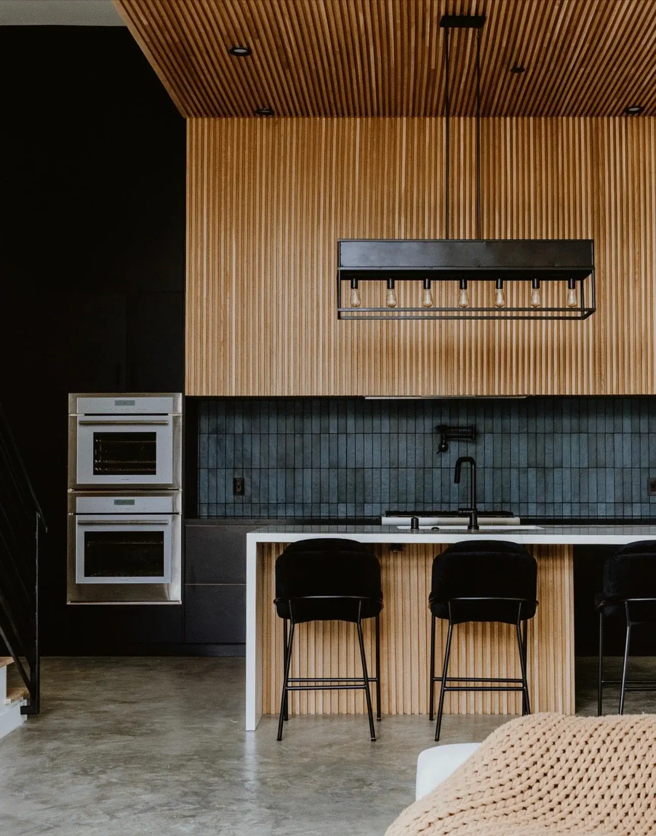 Modern kitchen with wood-paneled walls and ceiling, black backsplash, black chairs at a white countertop island, with an oven and built-in lighting fixtures.
