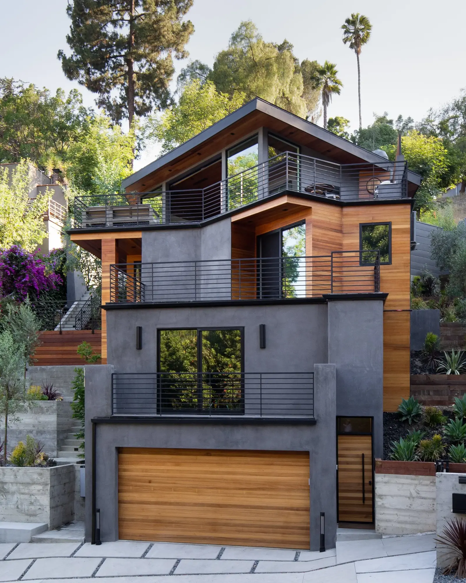 Modern multi-level hillside home in Northeast Los Angeles with warm wood siding, gray stucco, black metal railings, large windows, and multiple outdoor terraces. Modern multi-level hillside home in Northeast Los Angeles with warm wood siding, gray stucco, black metal railings, large windows, and multiple outdoor terraces.