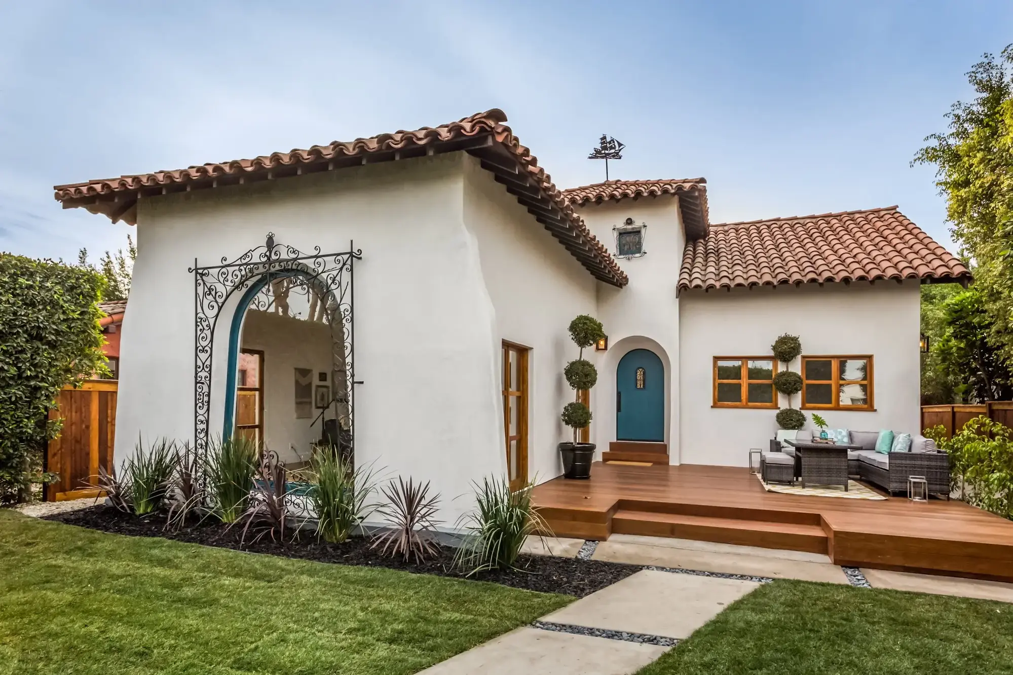 Spanish-inspired home exterior with white stucco, clay tile roof, arched windows, teal front door, warm wood deck, and wrought-iron detailing.