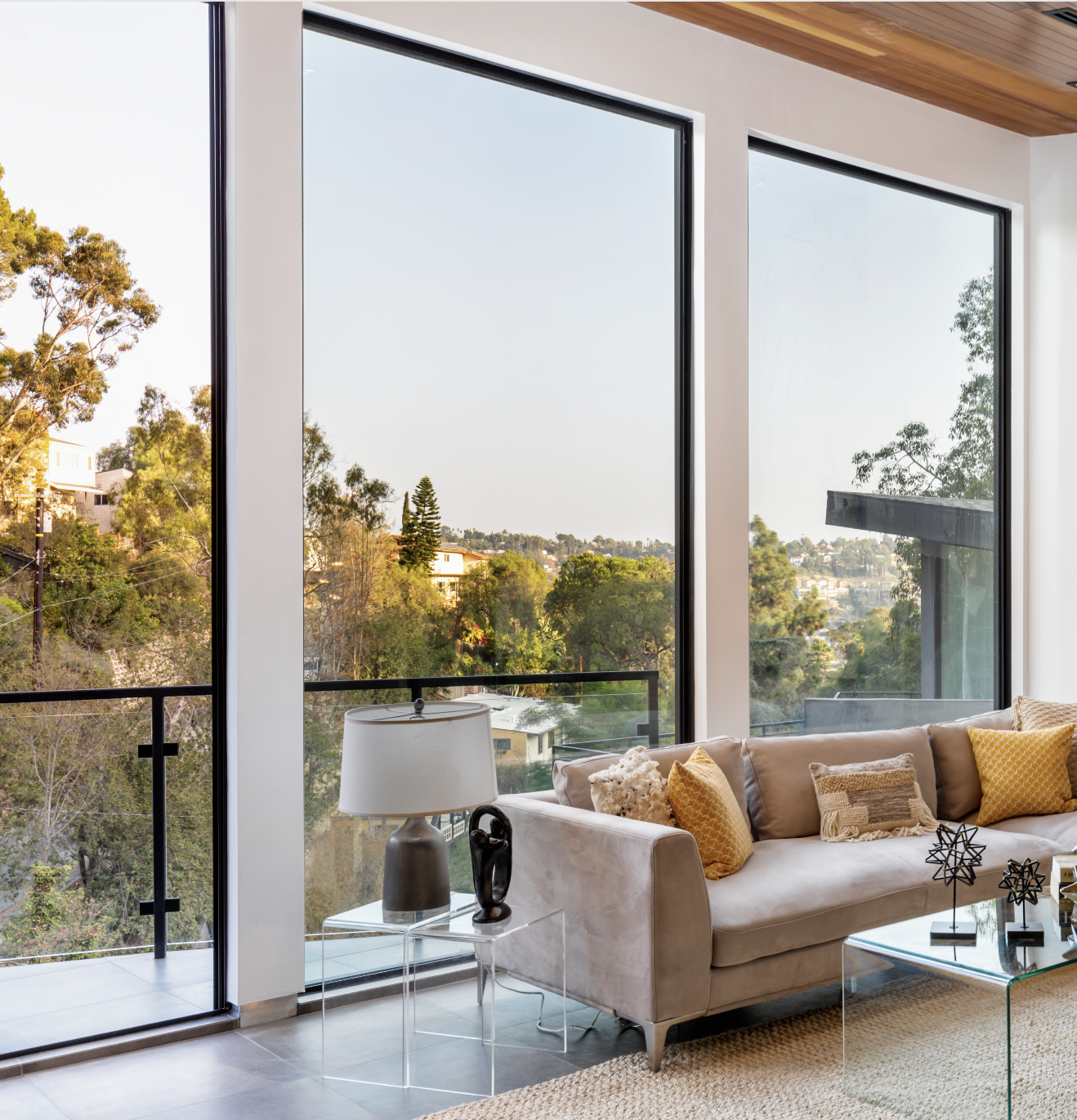 Modern living room with large floor-to-ceiling windows, a beige sofa with yellow and beige pillows, a transparent side table with a gray lamp and sculpture, and a glass coffee table, overlooking a lush green landscape.