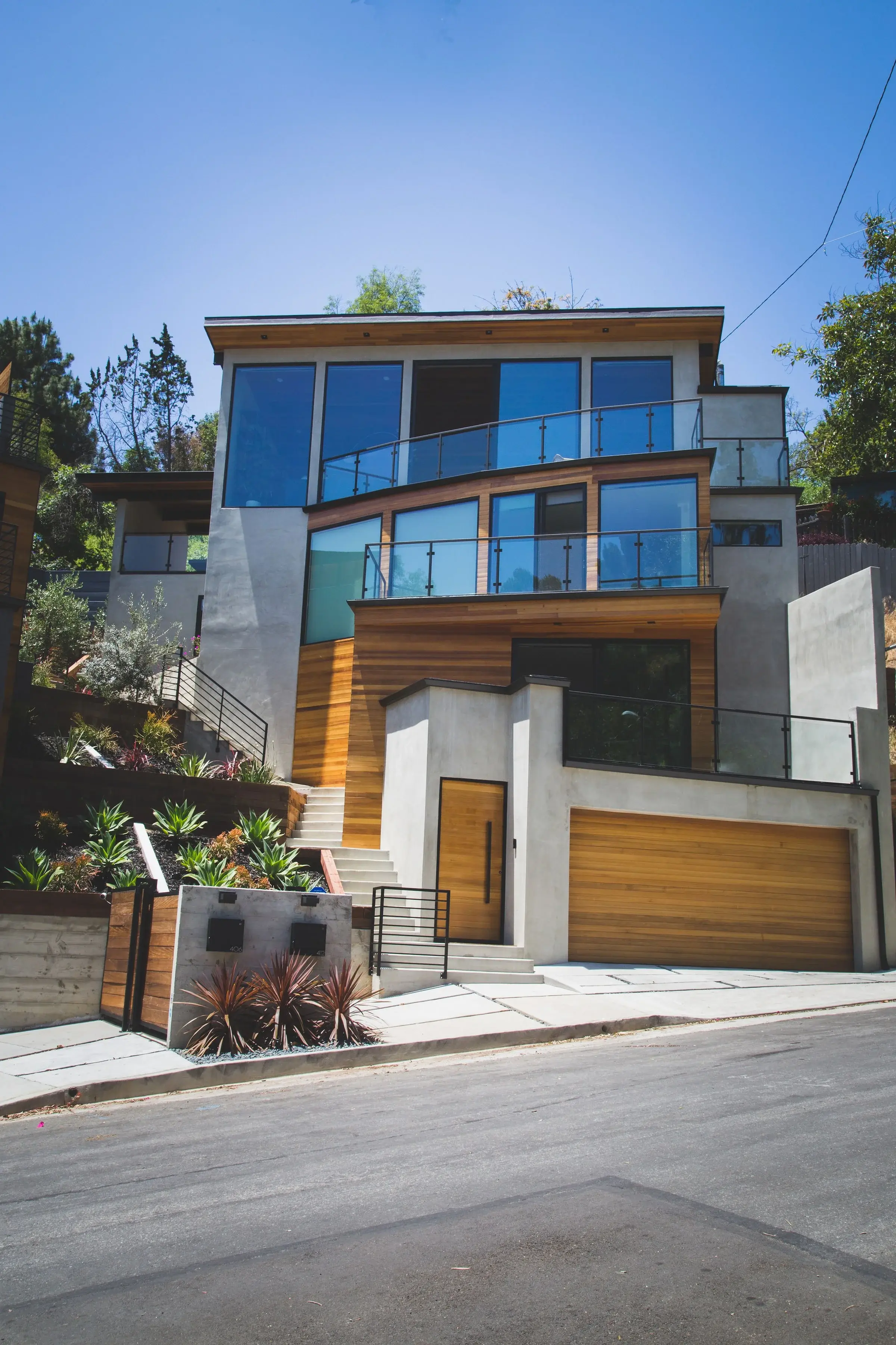 Modern multi-story house with large glass windows, wooden accents, and a garage, situated on a sloped street with landscaped yard.