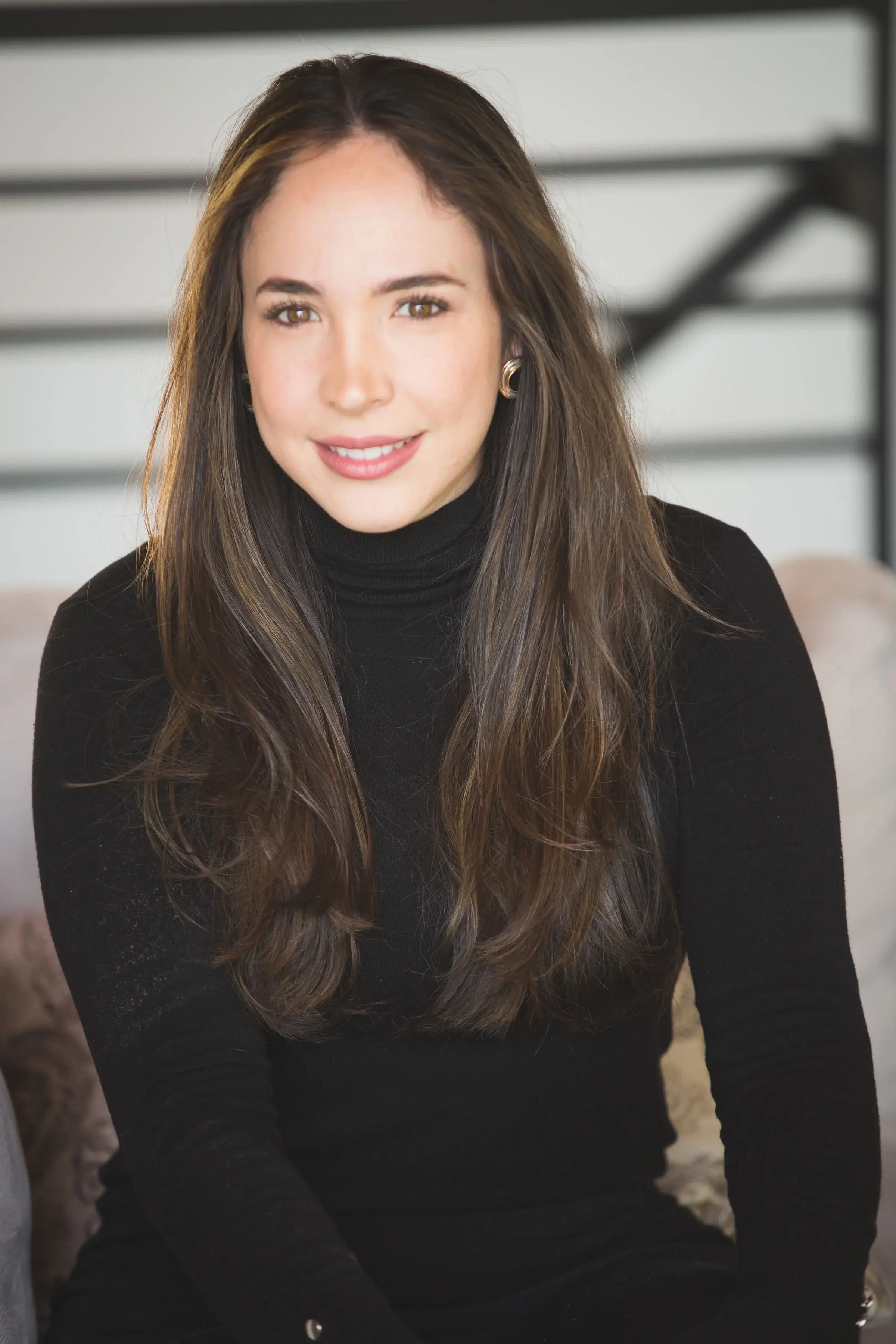 A woman with long brown hair, wearing a black turtleneck sweater, sitting indoors with a neutral background.