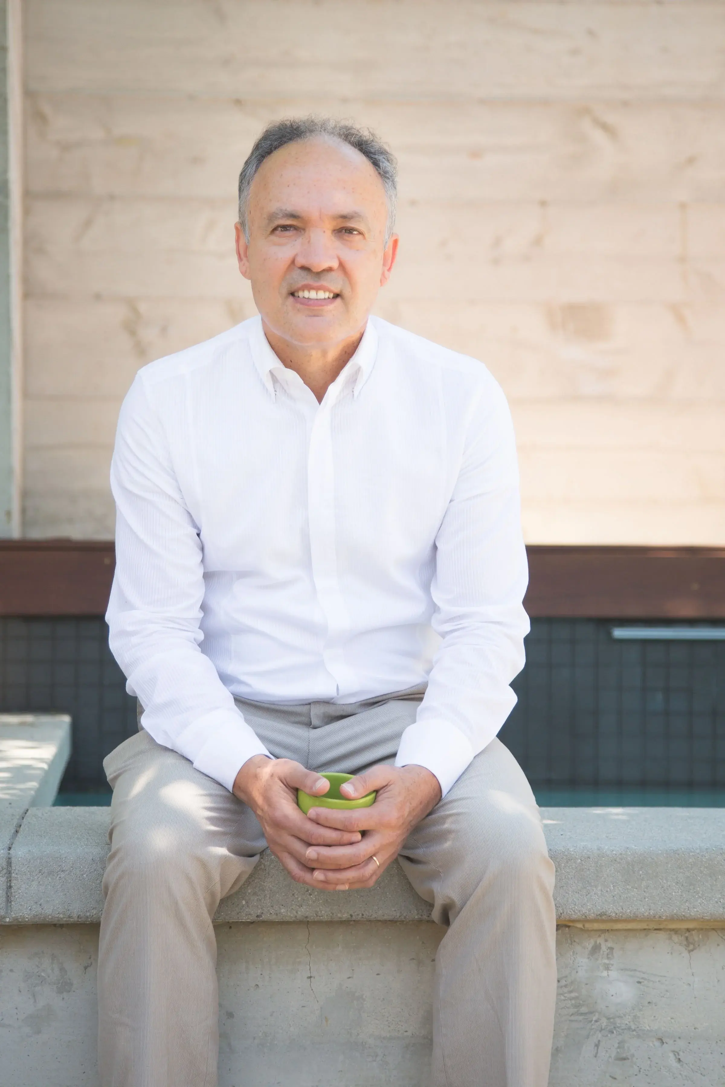 A middle-aged man with gray hair, wearing a white dress shirt and khaki pants, sitting outdoors on a concrete ledge holding a green smartphone.