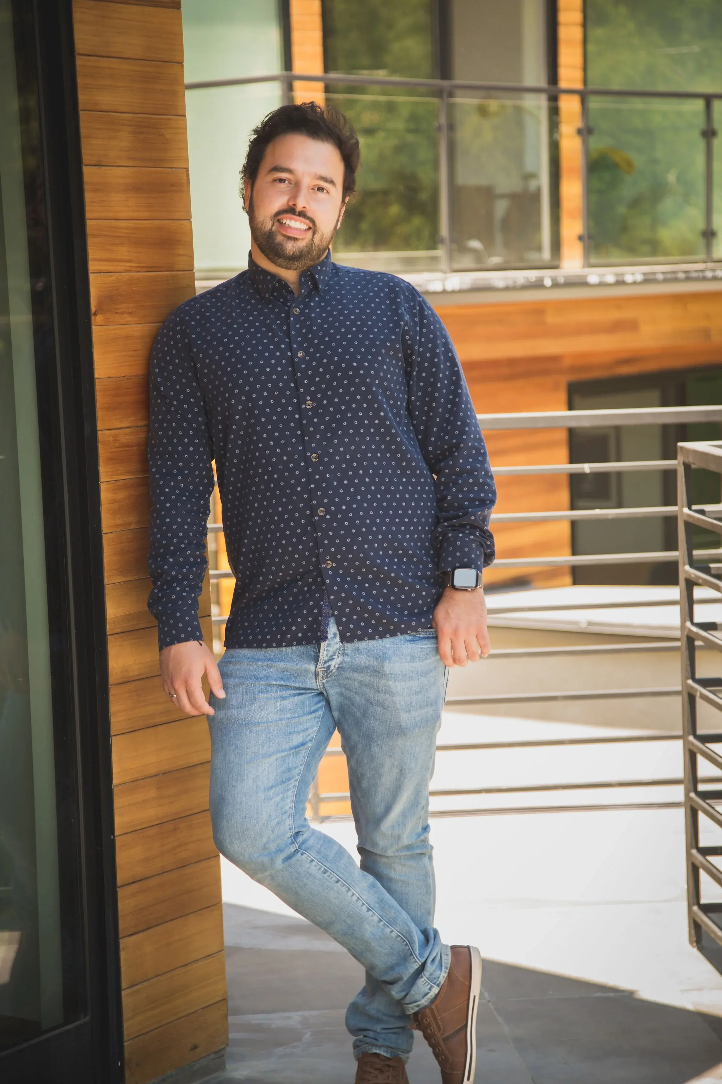 A man with dark hair and a beard smiling, standing outside on a balcony with wooden siding and metal railing, wearing a navy polka dot shirt, light blue jeans, and brown shoes.